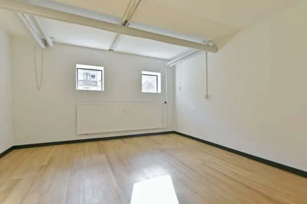 Lege kamer met houten vloer, witte muren, drie kleine ramen en zichtbare leidingen aan het plafond aan de Cornelis Dopperkade.